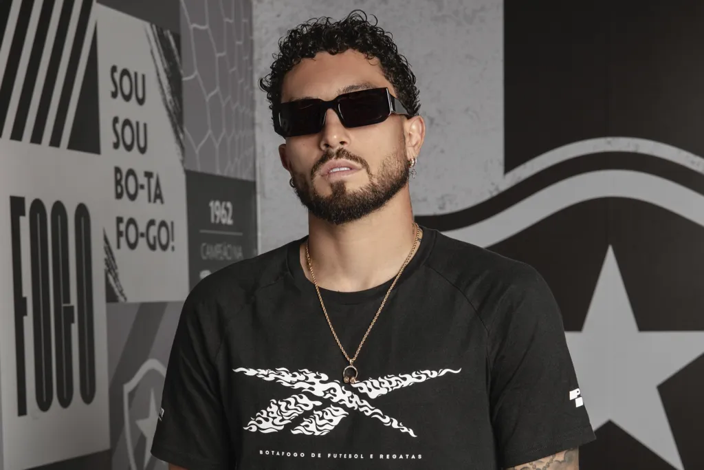 Jogador Alex Telles, do Botafogo, veste camiseta oficial e óculos Fuel Eyewear em frente ao painel do clube, durante ativação da parceria no Mundial de Clubes da FIFA 2025.