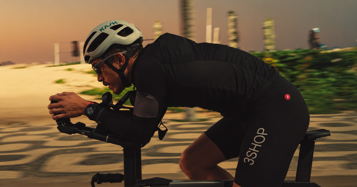 Homem praticando triatlo em uma bicicleta, focado e em posição aerodinâmica. Ele veste roupa preta de ciclismo, relógio esportivo, capacete e óculos escuros adequados para alta velocidade.