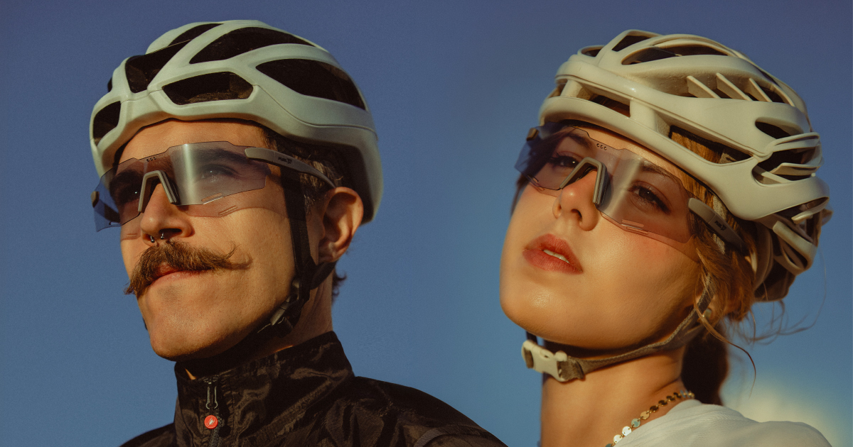 Um homem de bigode e uma mulher lado a lado contra um céu azul. Ambos usam capacetes de ciclismo e óculos esportivos transparentes e bem ajustados ao rosto