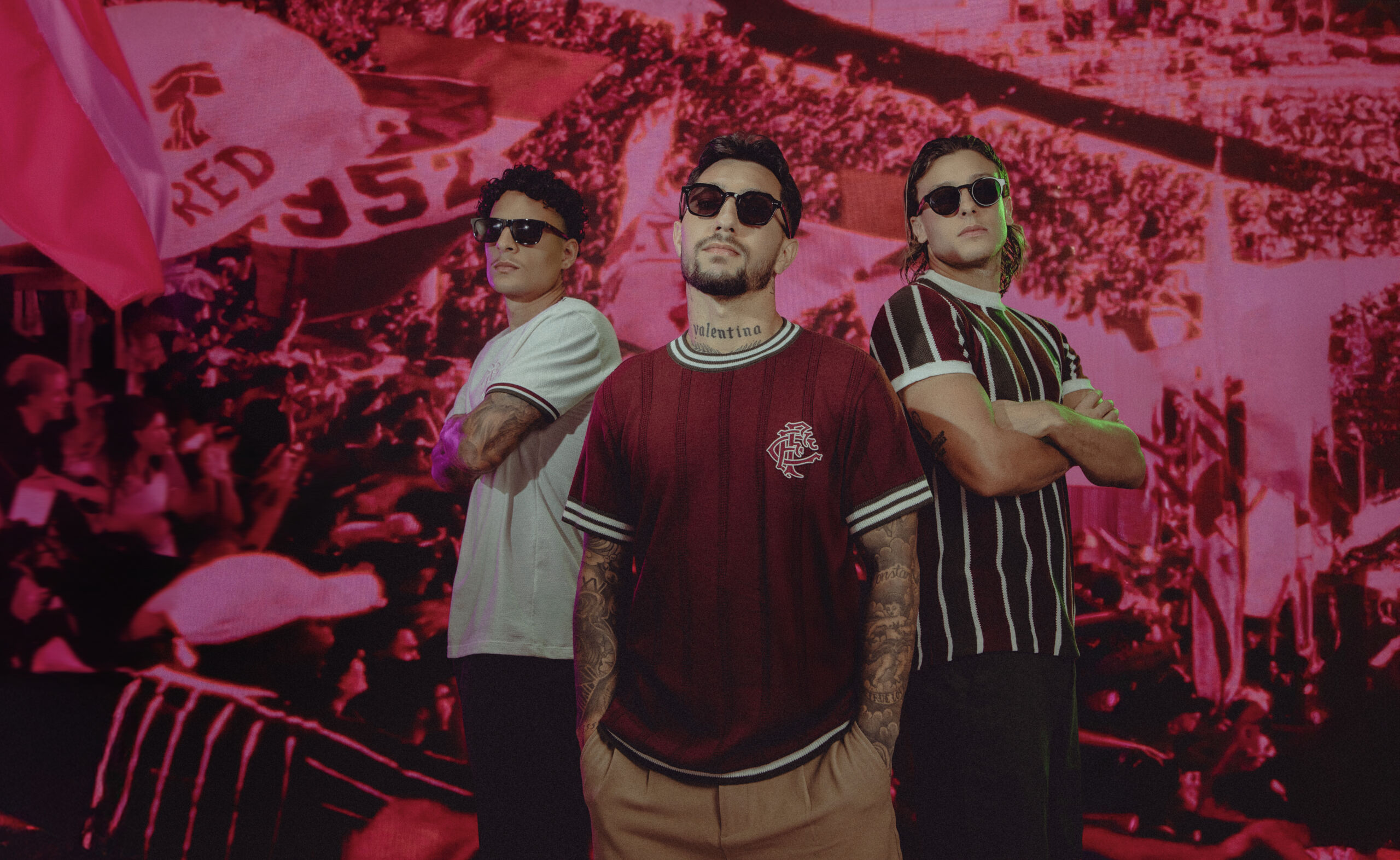 Três modelos em pé a usar óculos de sol e camisolas do Fluminense, com a bancada de um estádio cheia de adeptos ao fundo num tom avermelhado