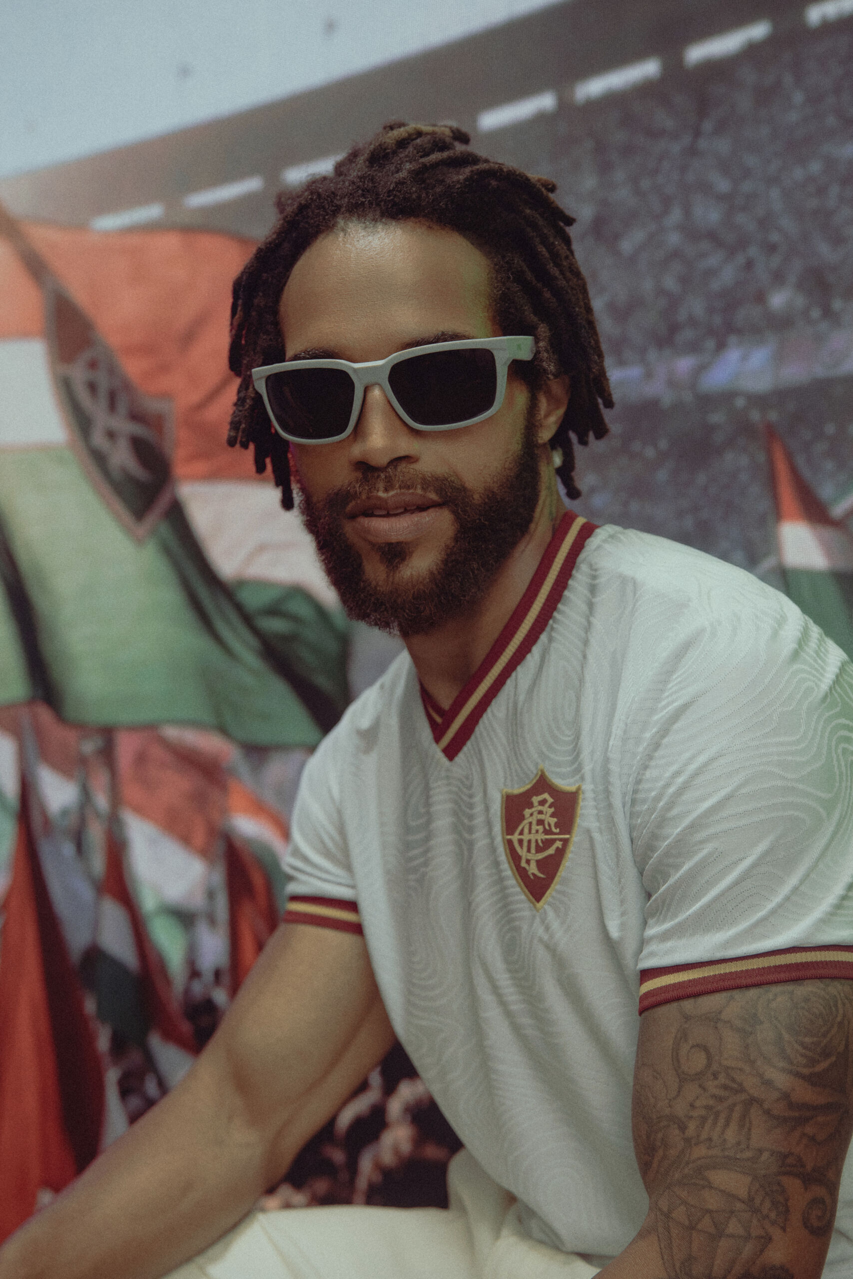 Modelo masculino de cabelo ondulado a usar óculos de sol hexagonais e camisola de tricot às riscas tricolores do Fluminense